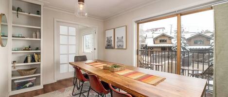 A dining area with a long wooden table, red chairs, a patterned rug, and a view of the neighborhood through large windows.