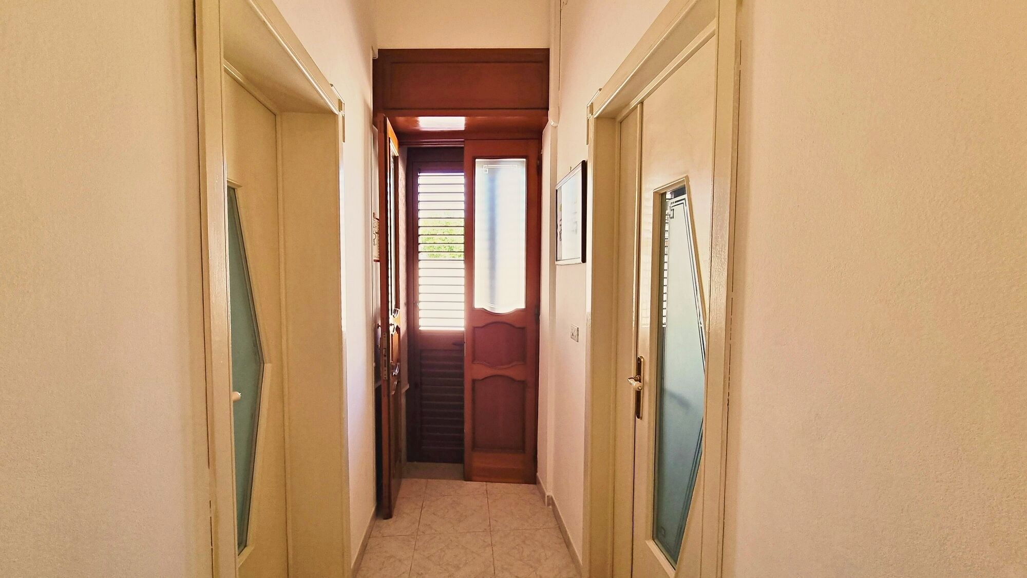 Ein schmaler Flur mit Türen an beiden Seiten, einem Fenster mit Jalousien und einer Tür mit kleinem Fenster.