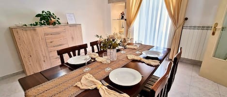 Ruokailutilassa on puinen pöytä, johon on katettu neljälle, sen vieressä on kaappi, jossa on kasvi, ja ikkunassa on verhot.