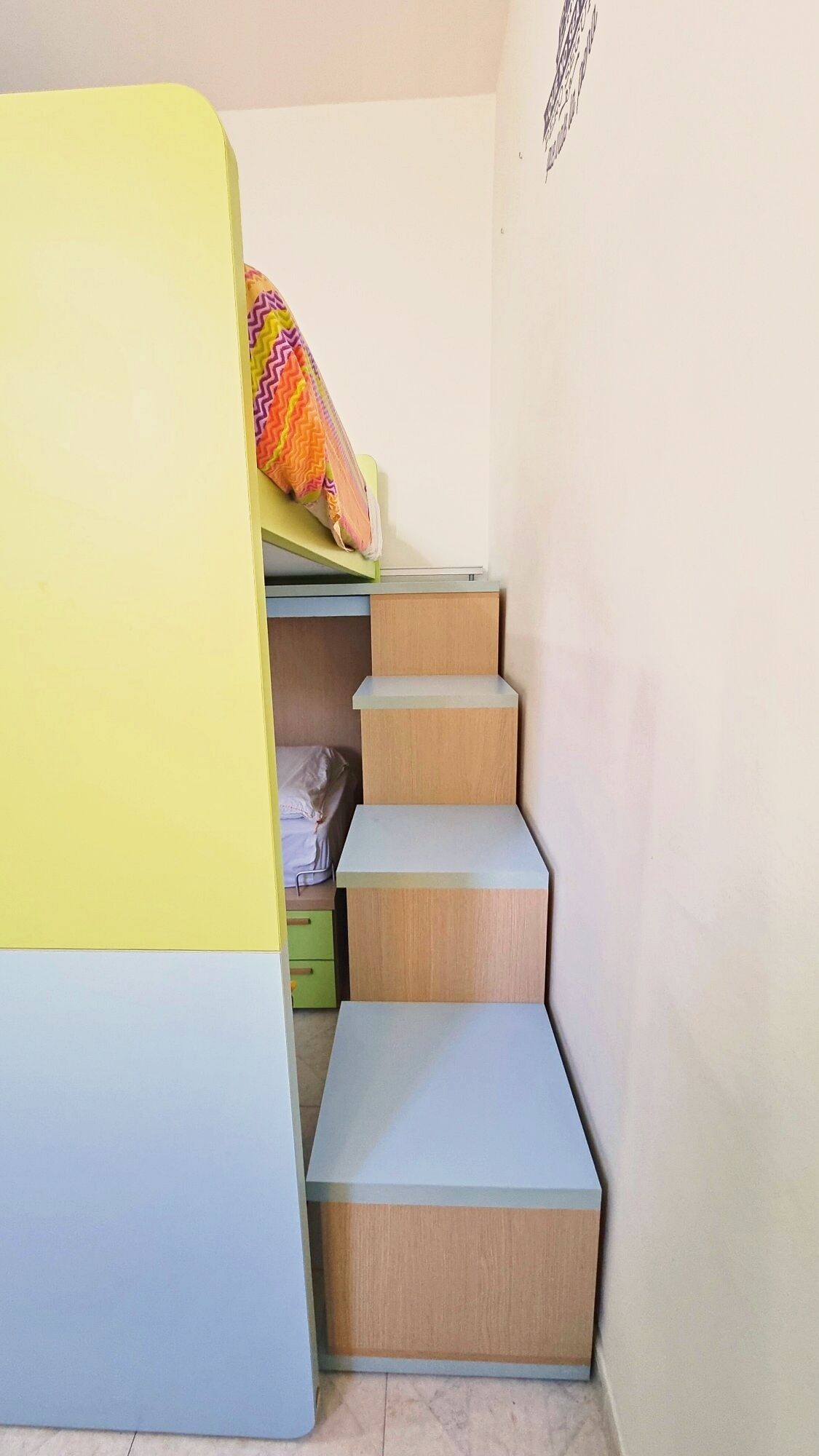 Una cama litera con un cojín de colores en zigzag, un escritorio pequeño y una cómoda con cajones.