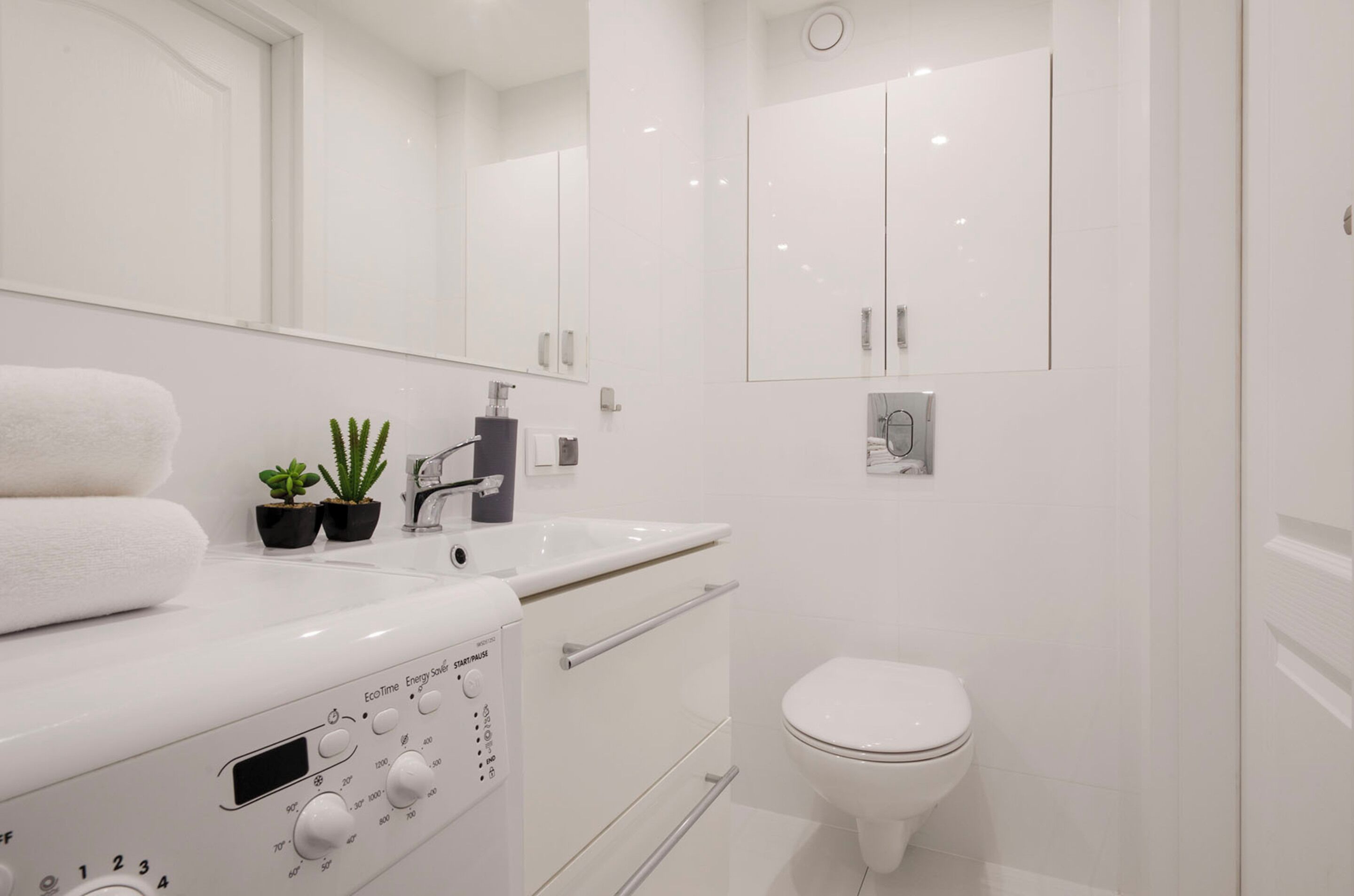 A modern bathroom with a white sink, a toilet, and a shower area. There are two potted plants on the sink counter.