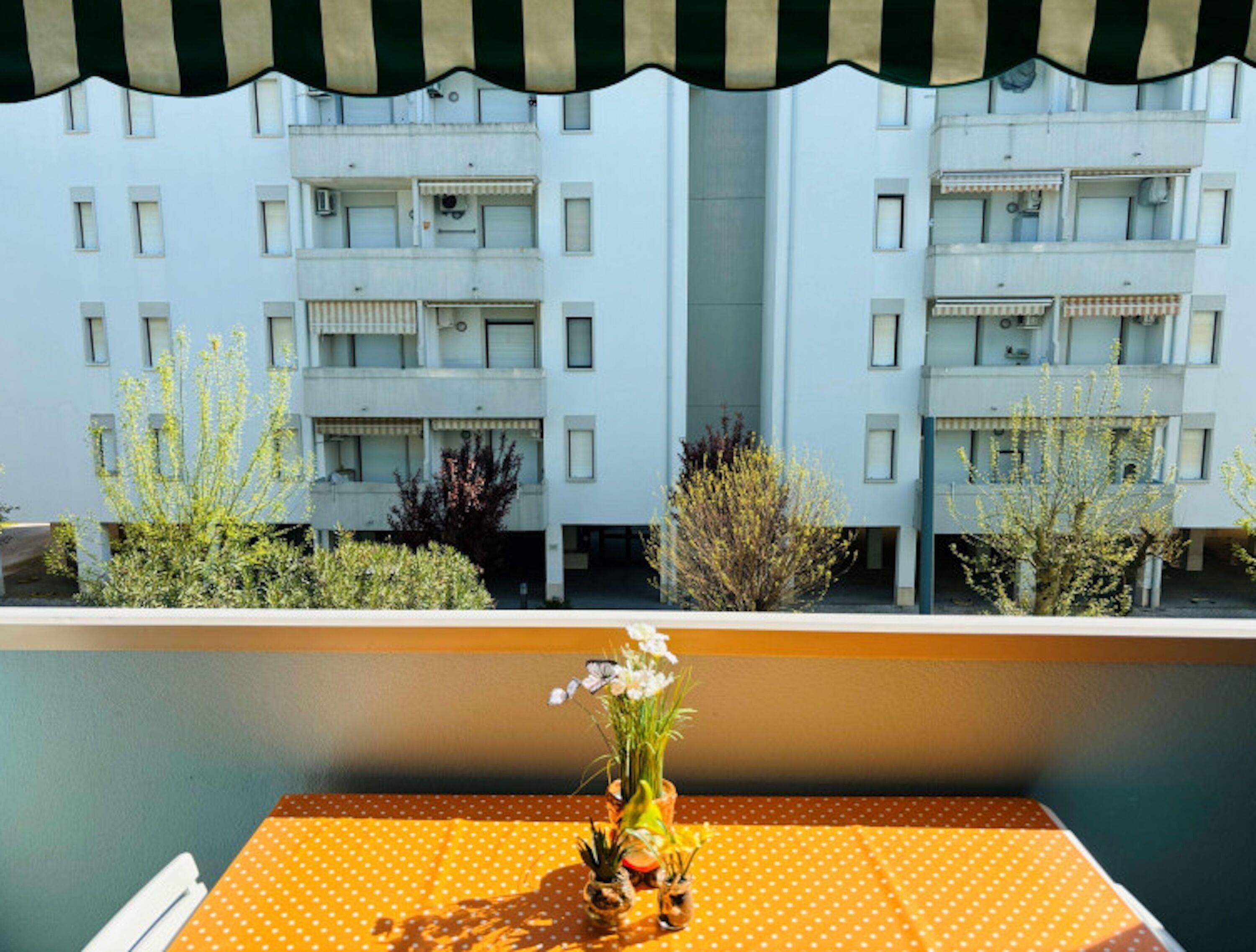 Un balcón con vista a un edificio de varios pisos, una mesita con un florero y un toldo a rayas.