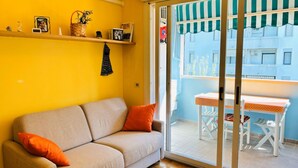 Ett vardagsrum med en beige soffa, orange kuddar, en liten hylla med diverse föremål och en skjutdörr i glas som leder ut till ett utomhusområde med ett bord och stolar.
