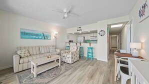 Sala de estar con sofá, mesa de centro y sillón estampado. Se observa una zona de cocina con refrigerador, microondas y taburetes de bar. Hay un ventilador de techo y un cuadro en la pared.