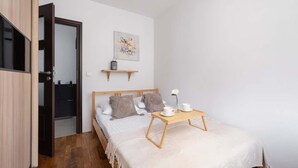 Un dormitorio con una cama, una mesita de noche de madera con una taza y un plato, un pequeño estante con un objeto decorativo, y una puerta que da a otra habitación.