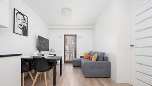 A modern living space with a dining area, a gray sofa with orange cushions, and a flat-screen TV on the wall. - Stylish Apartment in Praga | A Place to Work (Warszawa)