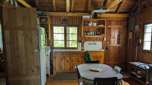 L’intérieur d’un chalet rustique est aménagé avec une table à manger en bois, des chaises, un réfrigérateur, un évier et une fenêtre donnant sur un paysage verdoyant.