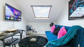 A compact living space with a TV mounted on the wall, a skylight, a blue sofa with vibrant pillows, a small round table with a glass top, and a black rug. - Cosy 1-bedroom apartment in beautiful location in London with WiFi and Netflix (London)
