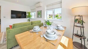 Uma sala de jantar com mesa de madeira posta para quatro pessoas, um sofá verde e uma TV fixada na parede.
