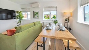 Uma sala de estar moderna com um sofá verde, uma mesa de jantar de madeira posta com pratos e copos, e uma televisão fixada na parede.