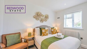 Quarto com cama, manta verde, cadeira de madeira, mesa de cabeceira e decoração na parede.