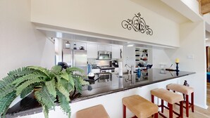 A modern kitchen with white cabinets, a stainless steel refrigerator, and a microwave. There is a bar area with stools and a large potted fern. - River City Resorts: RPS 104: River View Condo! Free Tubes Included! (New Braunfels)