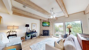 A cozy living room with a fireplace, a television mounted on a stone wall, a ceiling fan, and large windows offering a view of the outdoors. - River City Resorts: RPS 104: River View Condo! Free Tubes Included! (New Braunfels)