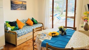 A cozy room with a sofa, a dining table, and a view of a garden. - Flat with patio overlooking the pool (Bibione)