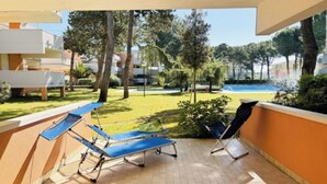 A covered outdoor area with a pool, lounge chairs, and a view of a green lawn and trees. - Flat with patio overlooking the pool (Bibione)