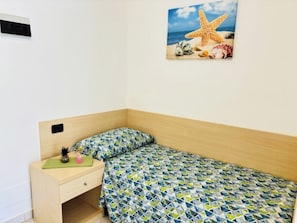 A bedroom with a wooden headboard, a patterned bedspread, a small bedside table with a green mat, and a wall-mounted air conditioning unit. - Modern and spacious studio in Bibione (Bibione Pineda)