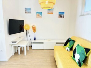 A modern living room with a flat-screen TV, a white sofa with green and black cushions, a white desk, a floor lamp, and wall art depicting beach scenes. - Beachside apartment with balcony (Bibione)