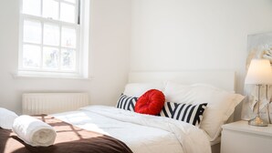 Un lit bien fait, avec une literie blanche et un plaid marron, un coussin rouge et un coussin à rayures. Il y a un radiateur blanc sous la fenêtre et une lampe sur la table de chevet.