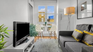 Ein modernes Wohnzimmer mit einem Flachbildfernseher, einem grauen Sofa mit gelben Kissen, einer Stehlampe und einem Essbereich mit Tisch und Stühlen.