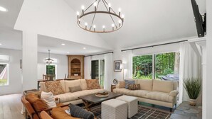 A spacious living room with a large sectional sofa, a coffee table, and a chandelier. There is a dining area with a table and chairs, and a view of the outdoors through large windows. - Charming Home in Harbor Springs Close to Town and Skiing (Harbor Springs)