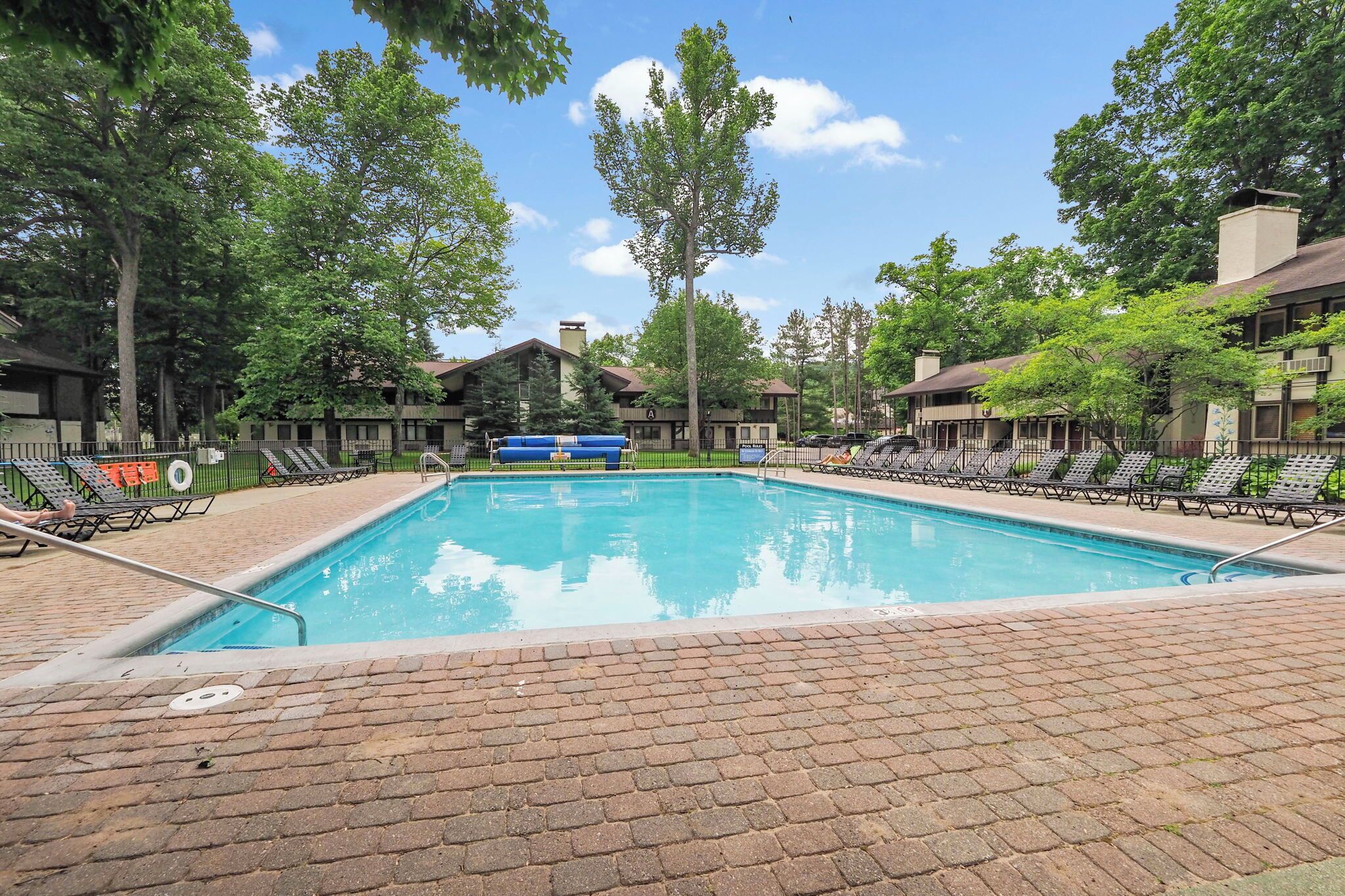 Une piscine rectangulaire bordée d’une terrasse en briques, entourée de chaises longues et d’arbres.