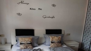 Een netjes opgemaakt bed met een gewatteerde spreidek en kussens, een nachtkastje met een lamp, en een muur met decoratieve tekst en hartjes.