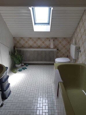 A bathroom with a skylight, white tiled floor, a green bathtub, a toilet, a radiator, and a potted plant. - Ferienwohnung Frank (Merzalben)