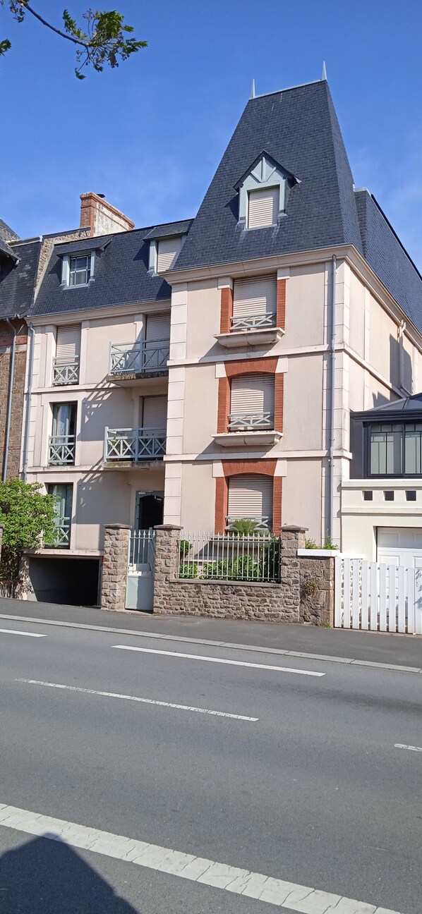 Un immeuble résidentiel à plusieurs étages, doté d’un toit en pente, d’un mur en pierre et d’une allée d’accès fermée par un portail.