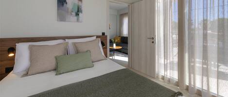 Camera d'albergo con un letto grande, una testiera in legno, una coperta verde e vista sull'esterno tramite tende trasparenti.