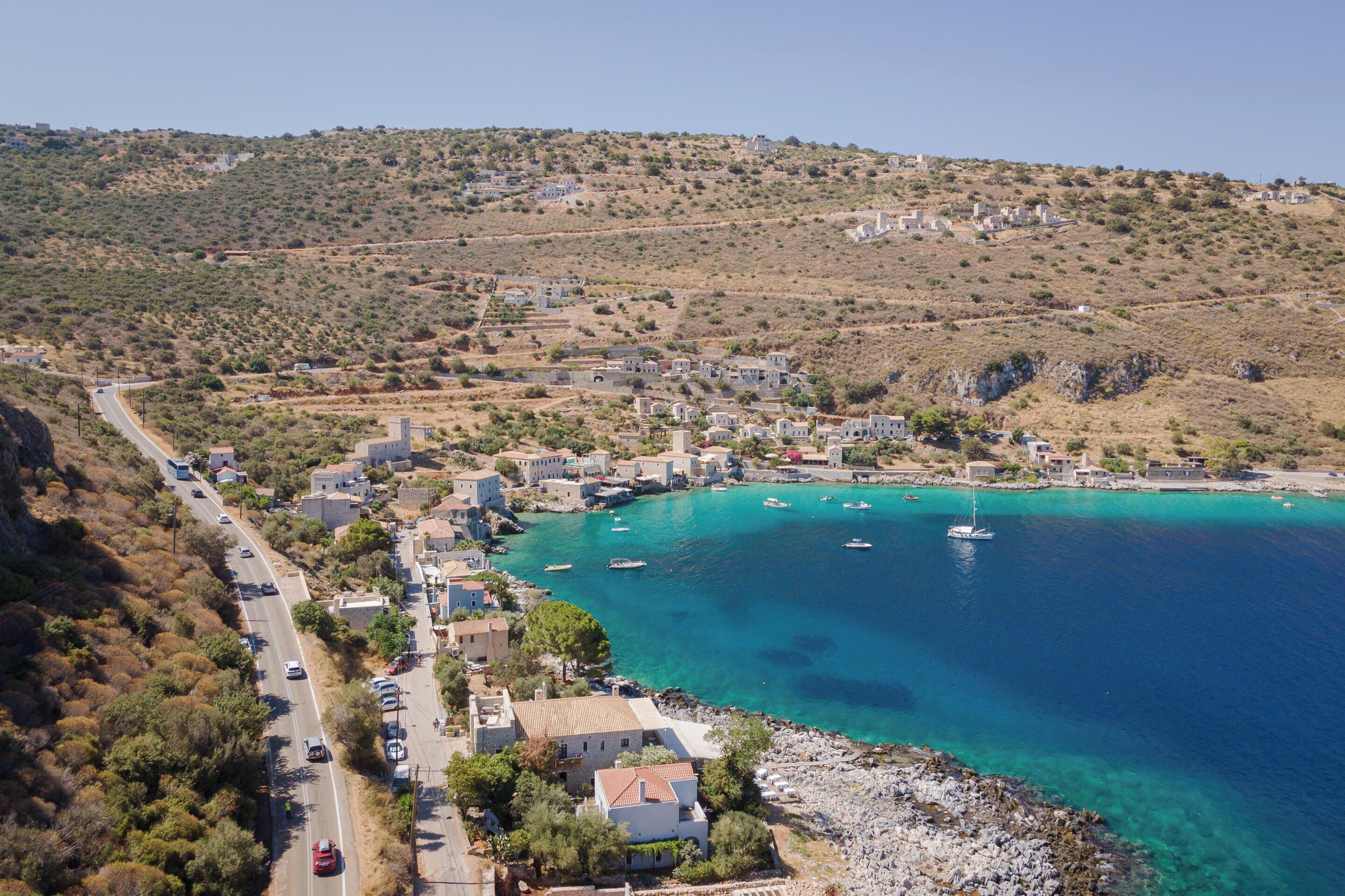 A coastal town with buildings along a curved road, a clear blue sea, and boats anchored nearby.