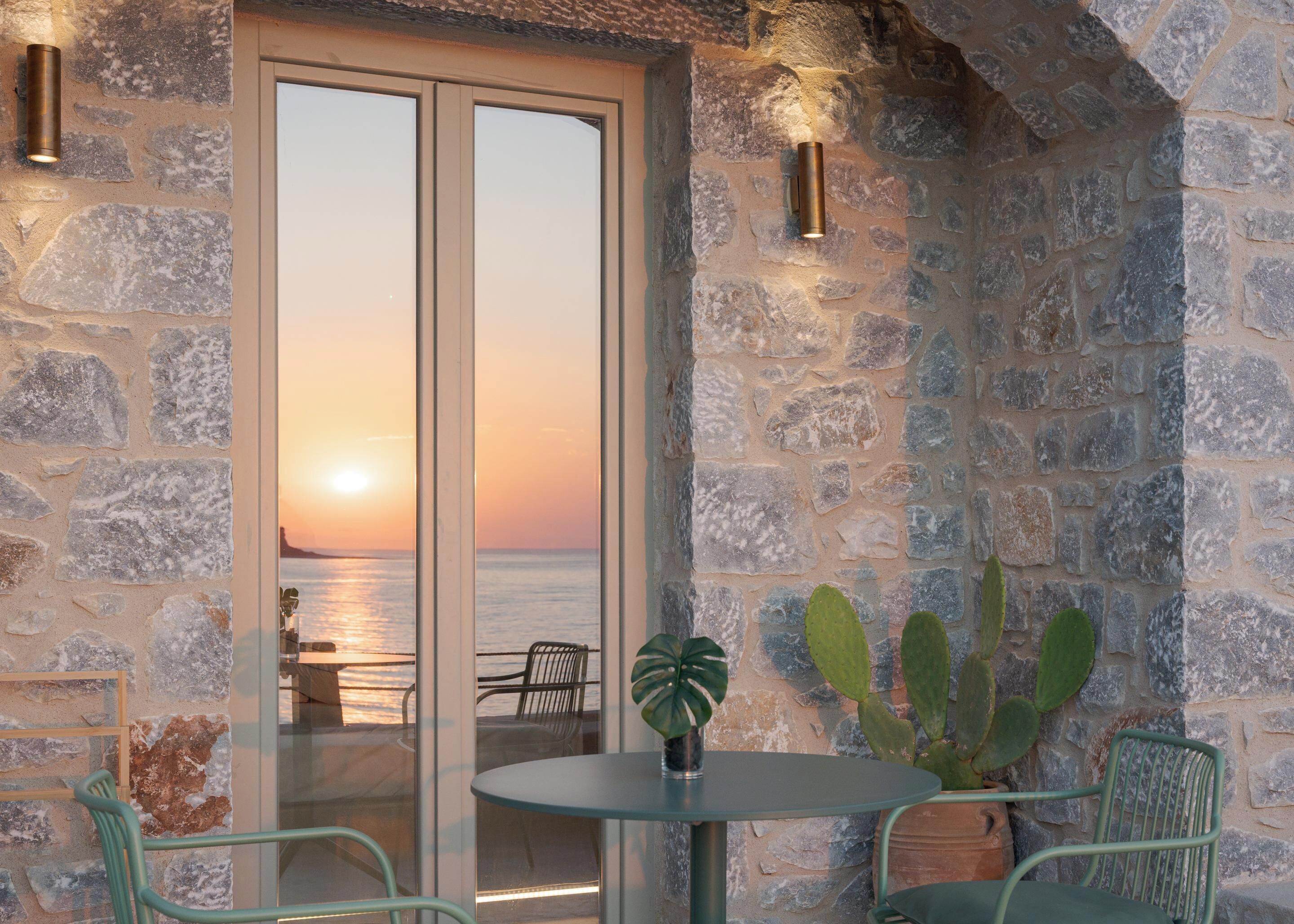 A stone wall with a glass door leading to a balcony overlooking a sunset over the sea.