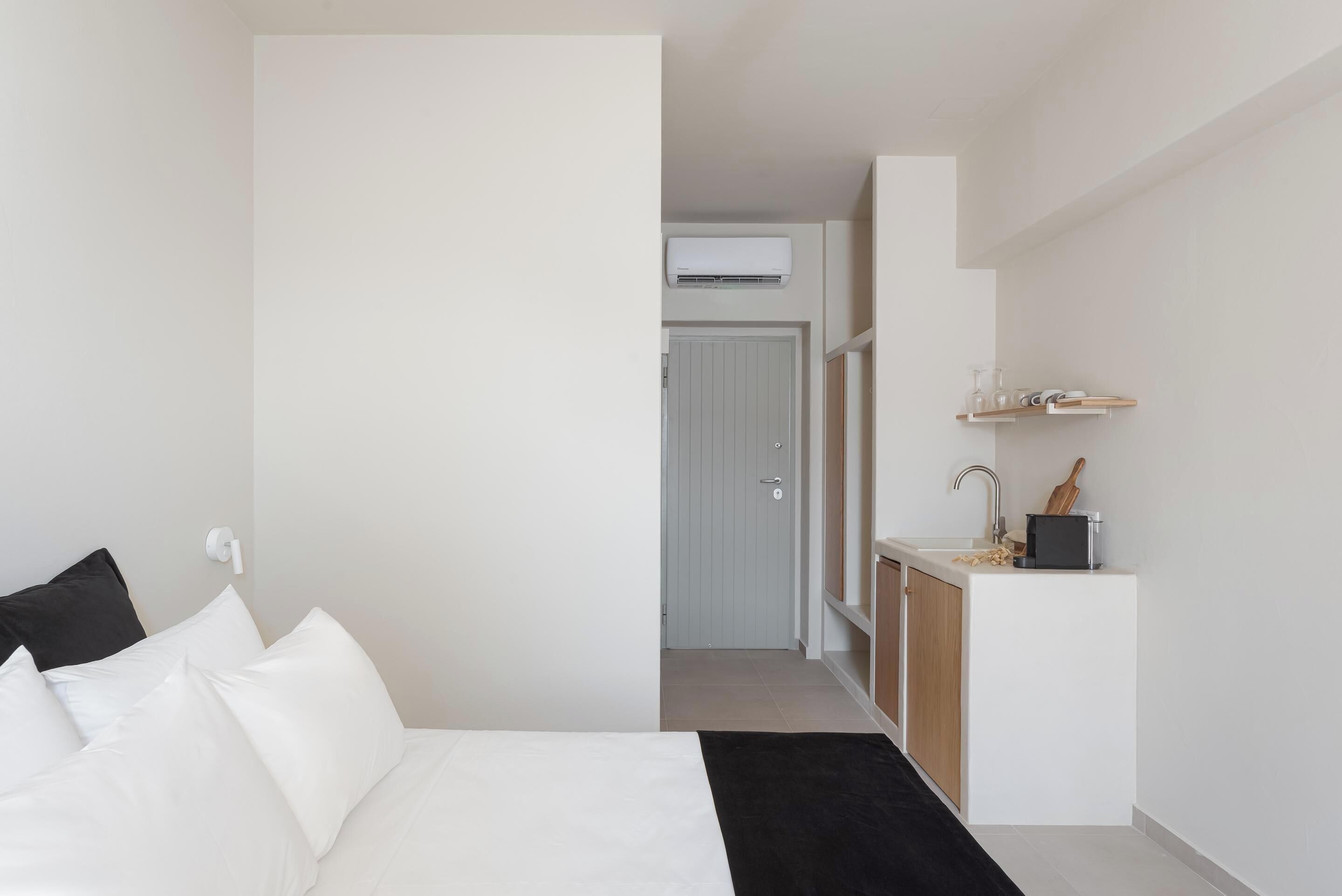 A modern hotel room with a bed, a small kitchenette, and a door leading to a bathroom.