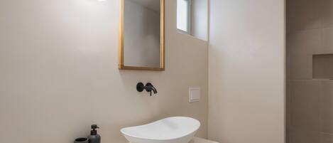 A modern bathroom with a white vessel sink, a round mirror, and a small window.