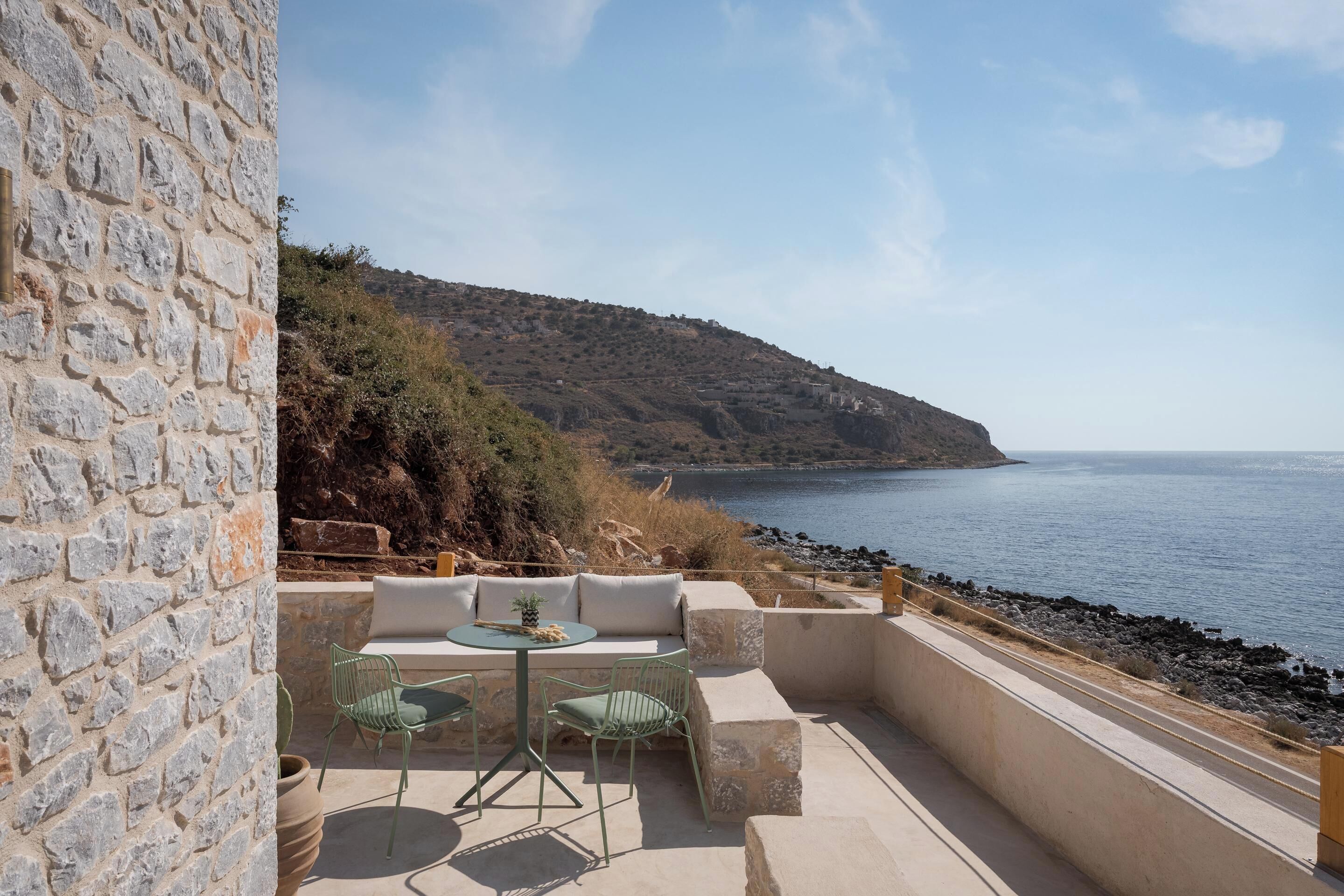 A stone wall, outdoor seating with a table and chairs, and a view of the sea and hills.