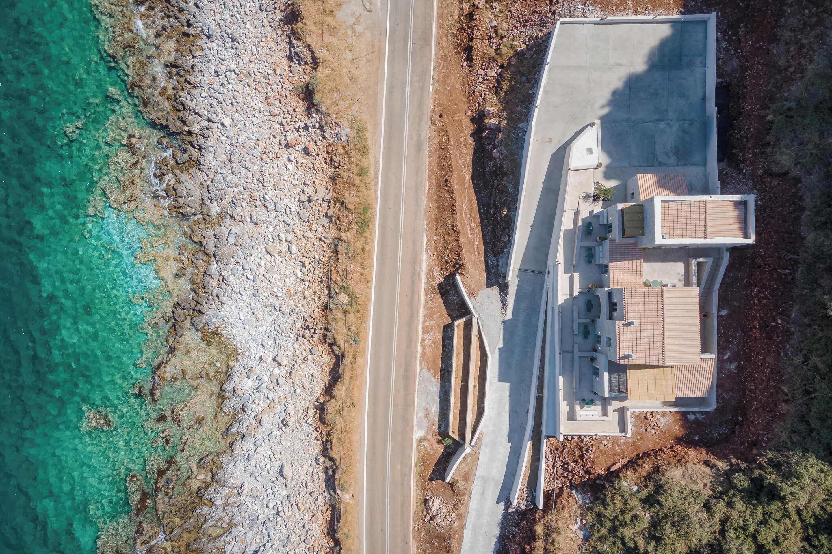 An aerial view of a coastal property with a paved road, a swimming pool, and a building with a tiled roof.