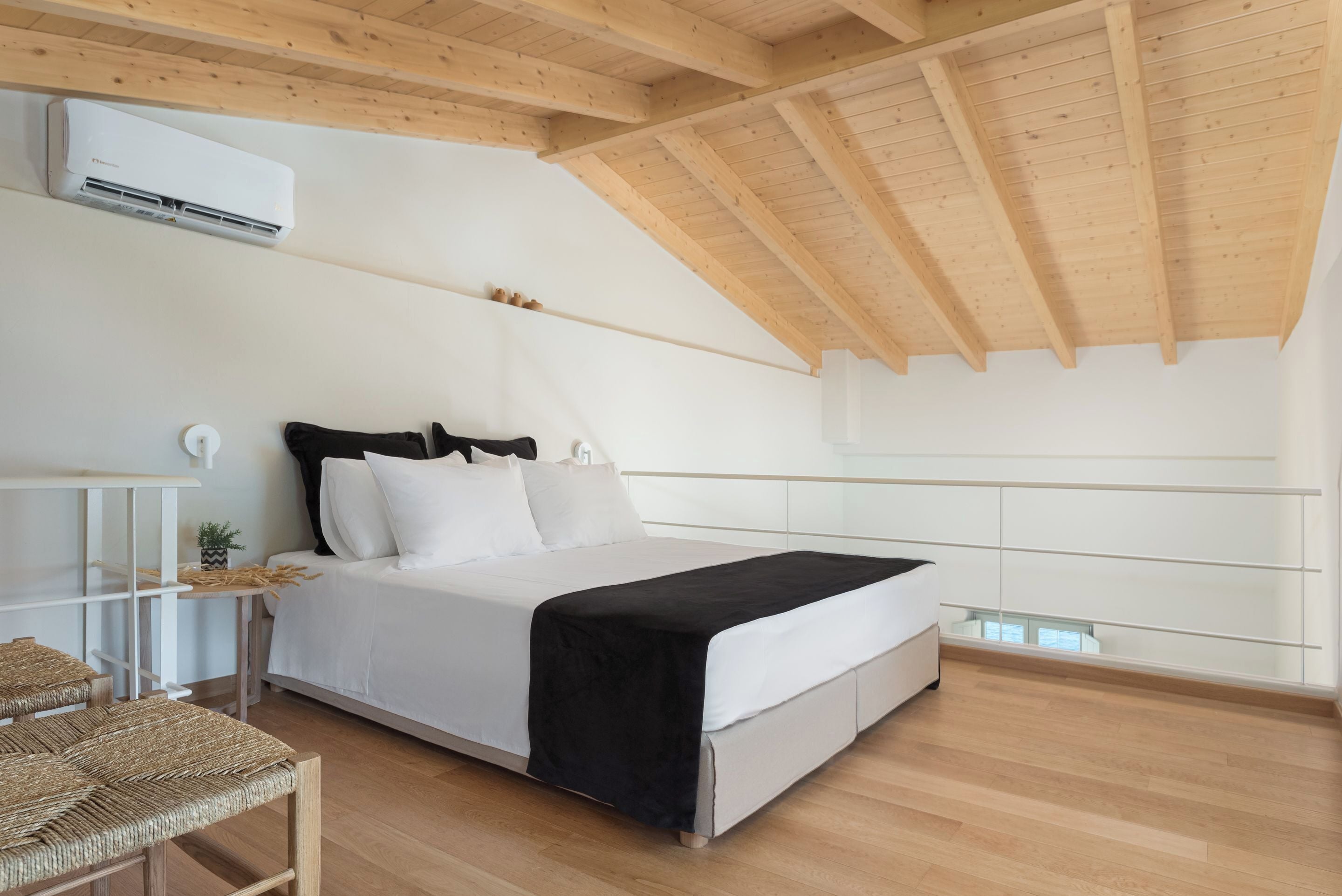 A bedroom with a sloped ceiling, a bed with white bedding and a black throw, a bedside table with a lamp, and a window with a view.