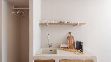 A kitchen with a white sink, wooden cabinets, a wooden cutting board, and a shelf with glassware and dishes.