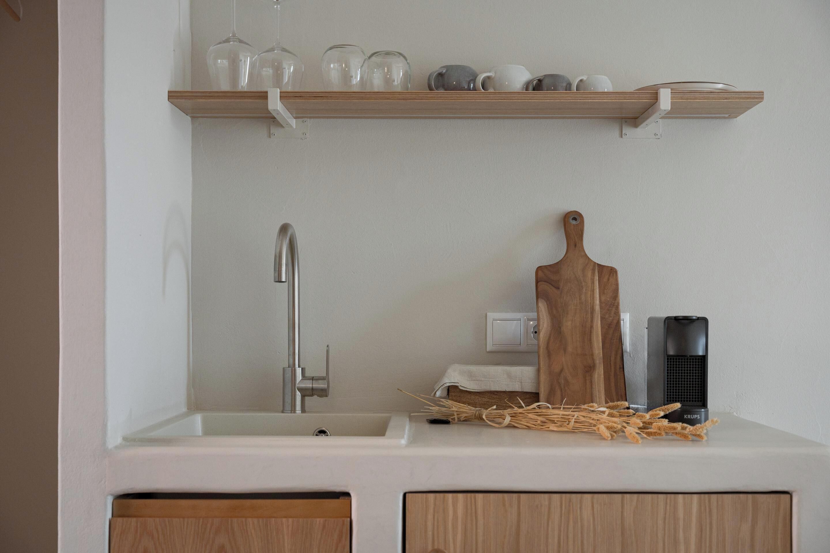 A kitchen with a white countertop, a wooden cutting board, a kettle, and a shelf above the sink holding various glassware.