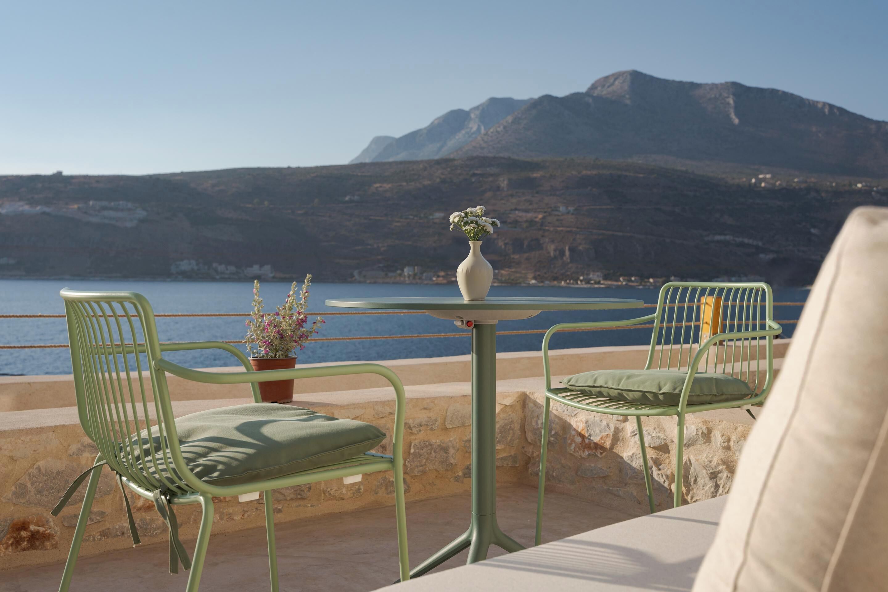 Una terraza con dos sillas metálicas verdes y una mesa, un florero con flores, y vista al mar y a las montañas.