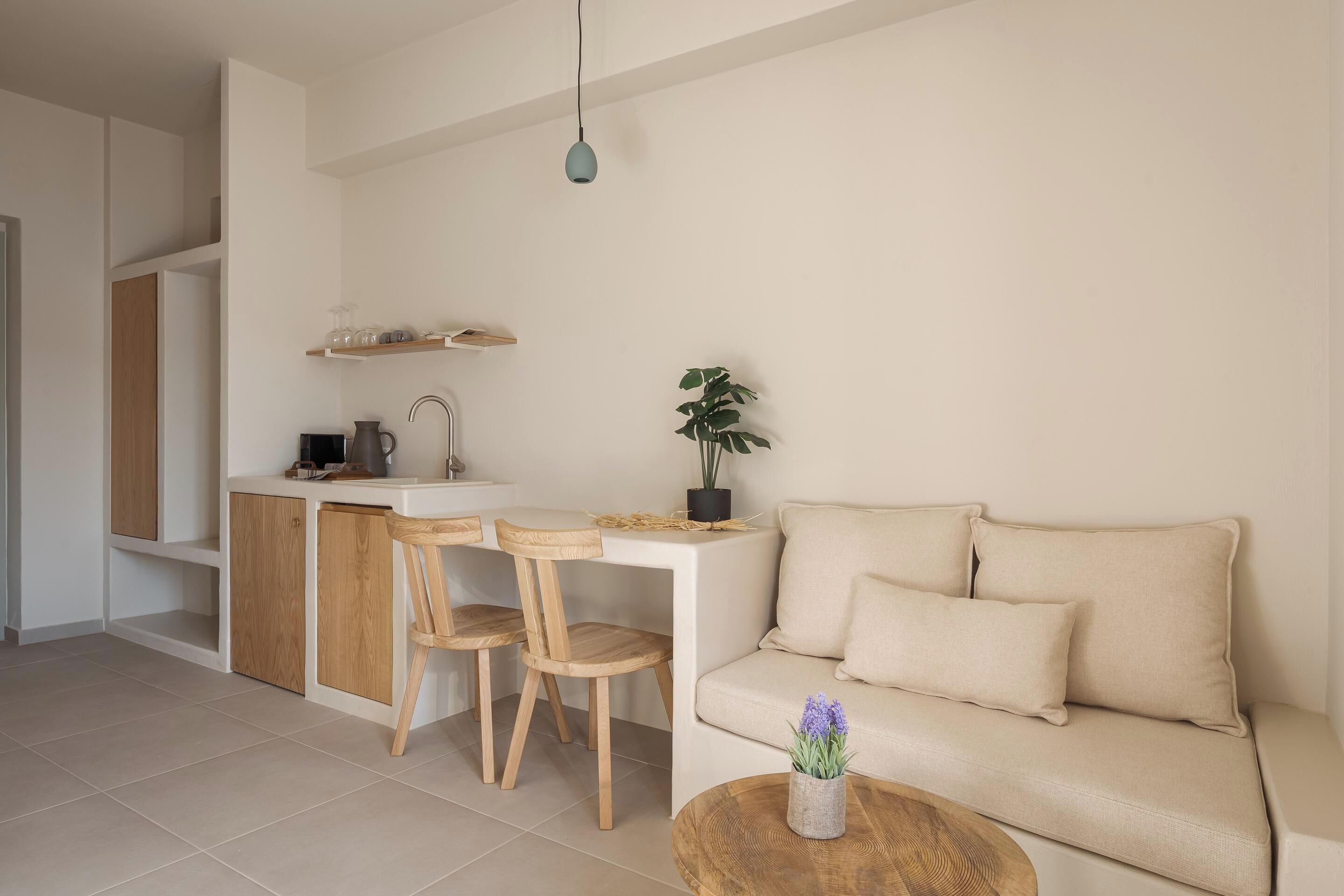 A modern kitchen with wooden cabinets, a countertop, and a sink. There is a dining area with two wooden chairs and a small table. A beige sofa is placed against the wall, and a potted plant is on the countertop.
