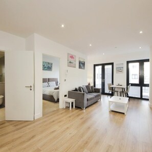 A modern living room with a grey sofa, a white coffee table, and a small white side table. There is a bed with a grey headboard in the adjacent room. The room has wooden flooring and large windows with black frames. - Modern Comfort: Seven Stays Broadoaks (Solihull)