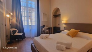 Quarto de hotel com uma cama grande, uma secretária e uma cadeira. Há uma janela com cortinas, um candeeiro fixado na parede e uma cabeceira.