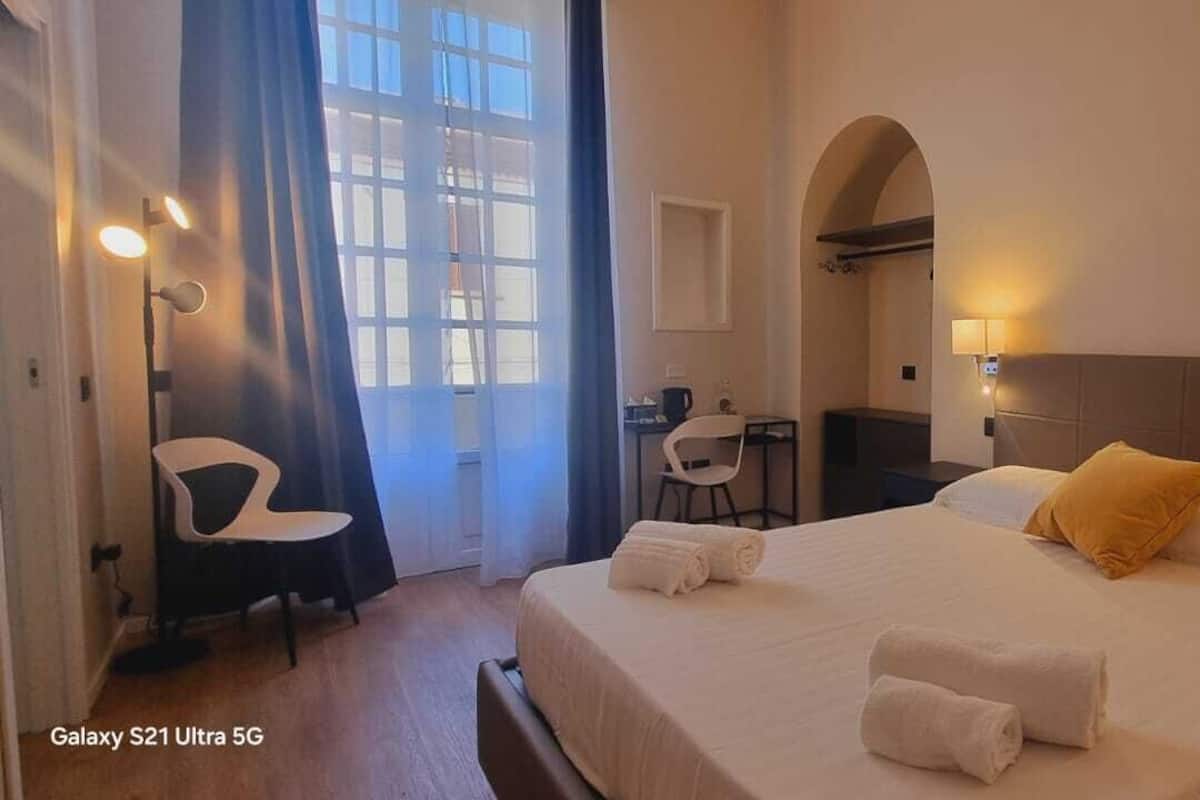 A hotel room with a large bed, a desk, and a chair. There is a window with curtains, a wall-mounted lamp, and a headboard.