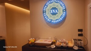Uma área de recepção de hotel com um logotipo na parede, uma mesa de buffet com comida e bebidas, e um celular Galaxy S21 Ultra 5G.