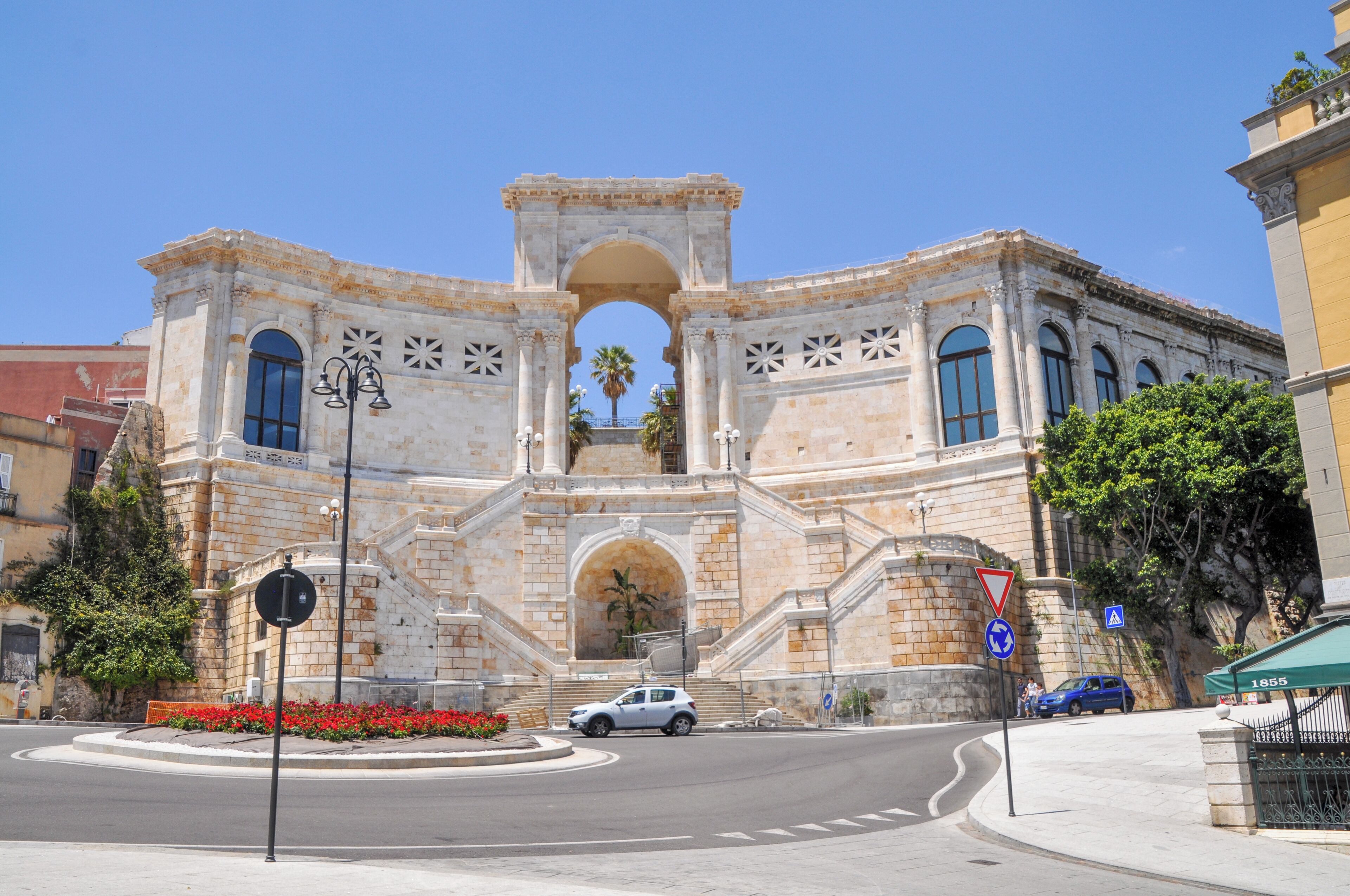 A historic building with a central archway, a roundabout, and a blue car parked in front.