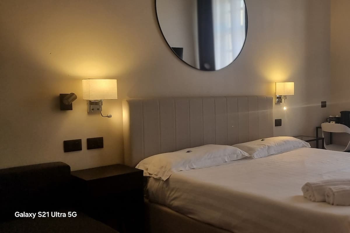 A hotel room with a large bed, two bedside lamps, a round mirror, a desk with a chair, and a window with curtains.