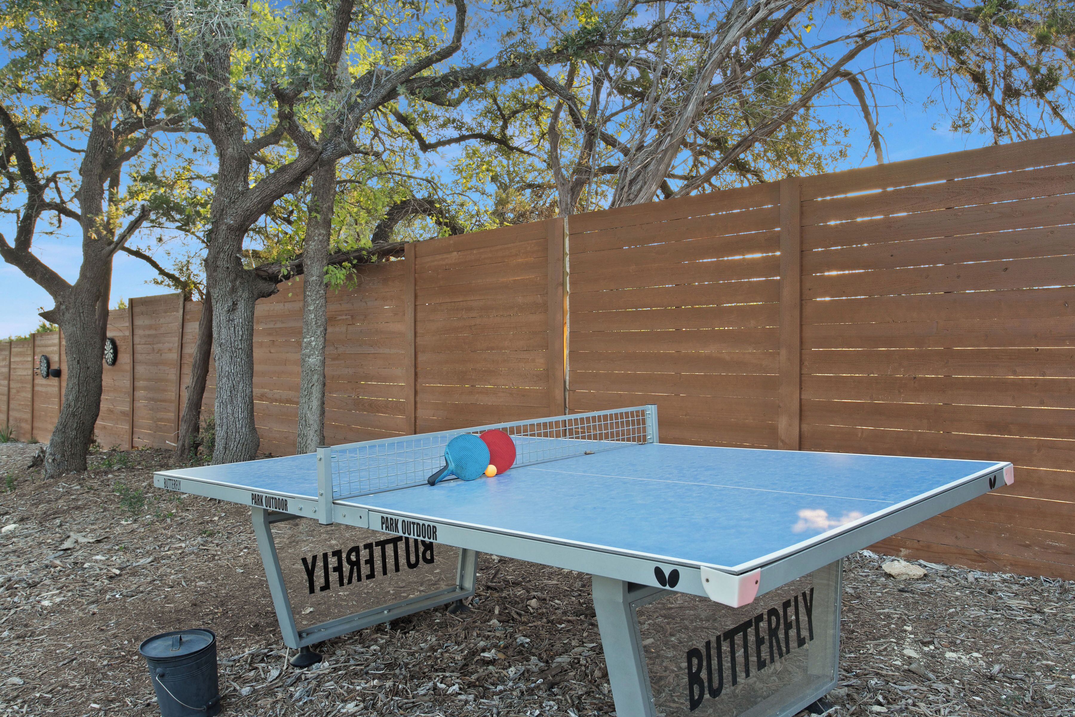 A table tennis table outdoors with a wooden fence and trees in the background.