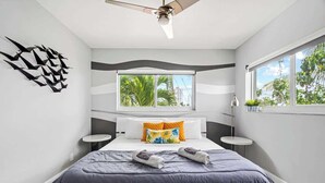 A modern bedroom with a large bed, a window with a view of palm trees, a ceiling fan, and a decorative wall piece. - Waterfront Retreat with Pool & Steps to Beach (Pompano Beach)