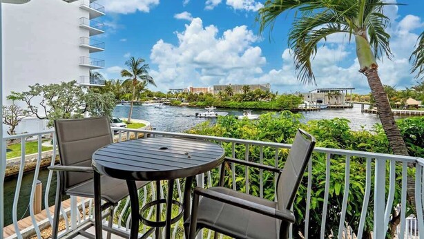 A balcony with a table and chairs overlooking a river, surrounded by greenery and a clear sky. - Waterfront Retreat with Pool & Steps to Beach (Pompano Beach)