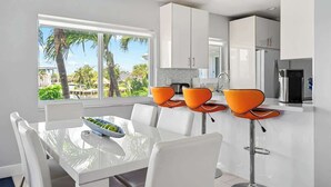 A modern kitchen with white cabinets, a stainless steel refrigerator, and a dining area with white chairs and a glass table. - Waterfront Retreat with Pool & Steps to Beach (Pompano Beach)